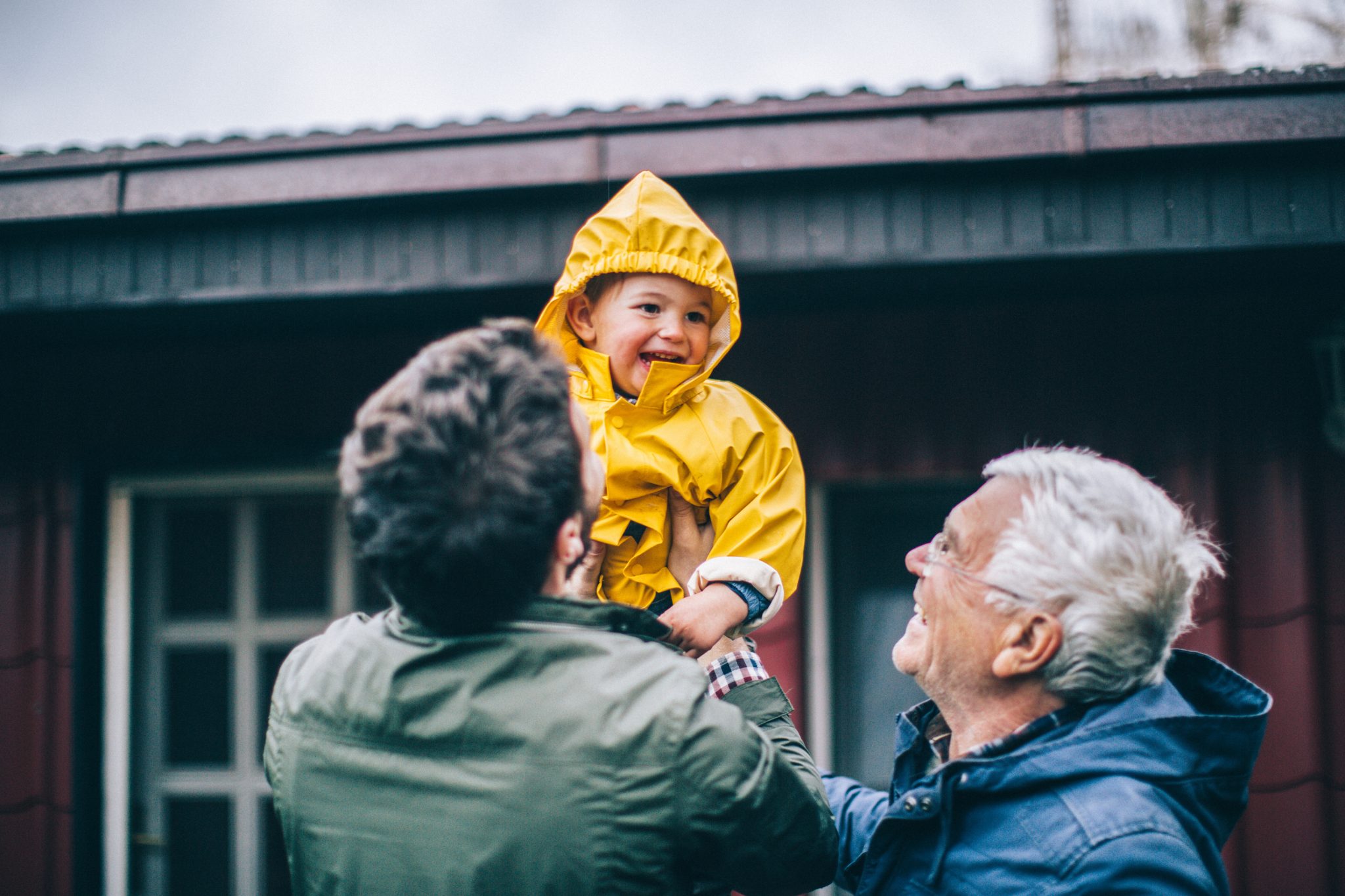 A young boy in a yellow raincoat being thrown in the air by his father and grandfather